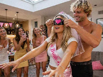 A fun hens party game where a blindfolded bride wearing a sash reaches forward while a topless waiter in black pants helps guide the activity. A group of women in summer outfits laugh, film, and cheer in a bright Airbnb setting, capturing a playful and interactive hens party game experience with LuckyLadds.