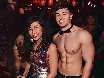 A topless waiter wearing black pants and a bow tie posing with a guest at a lively nightclub-style hens party. The scene features colourful lighting, a crowded dancefloor, and a fun, high-energy party atmosphere, showcasing the entertainment experience offered by LuckyLadds.
